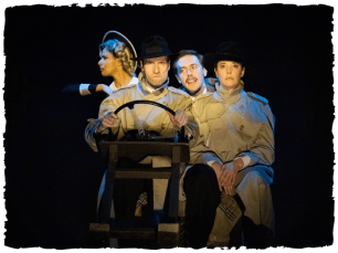 4 performers in coats drive behind a steering wheel