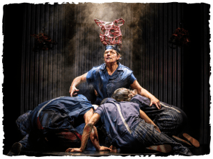 A performer wearing blue fabrics and a hat made of wire shaped like a pigs head is surrounded by other performers in front of a metal background