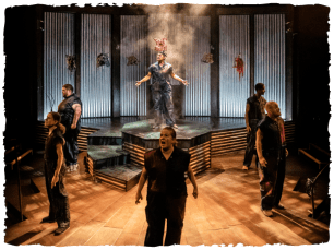 Performers are spread across the stage speaking at once, with one on a pedistal in the middle with a pig mask above their head