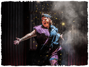 A performer is showered with gold glitter as they dance around in red and blue fabric clothes and a horn on their head