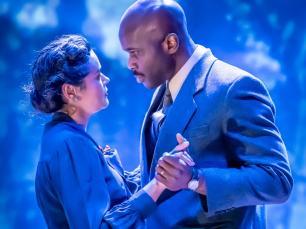 Two performers in smart outfits dancing together in front of blue backgorund