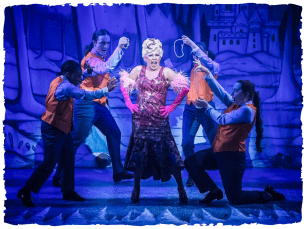 Lucy Benjamin (Baroness Hard-Up) sings in a red flowery dress surrounded by an ensemble in florescent orange jackets