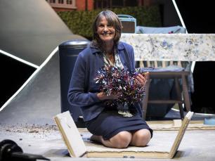 Gwyneth Strong (Vera) kneels on the floor. A hatch is open in the floor and she has pulled up a bundle of  multicoloured tinsel into her lap. She smiles out into the audience.