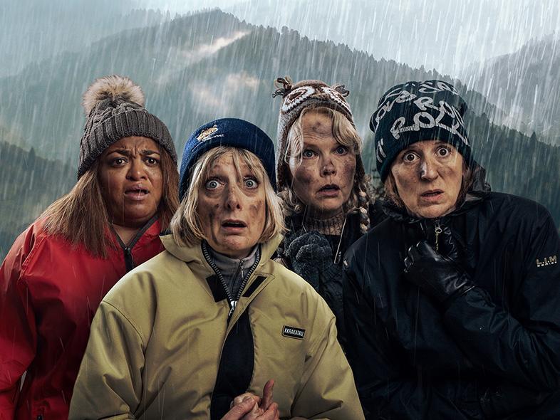 A group of four women in outdoor weather gear stand outside in a hilly area.