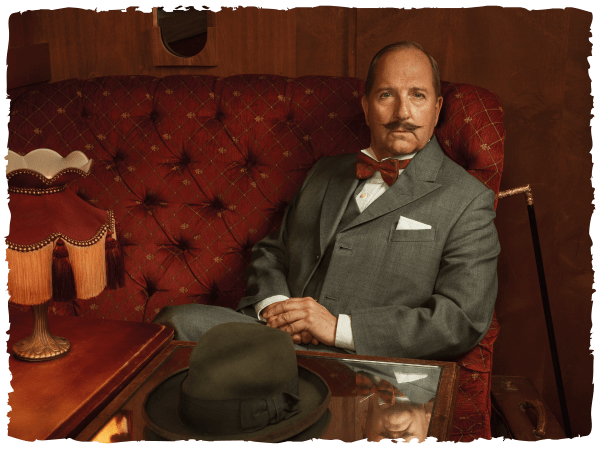 Michael Mahoney as Poirot in a grey suit, wiht red bow tie and iconic large mustache