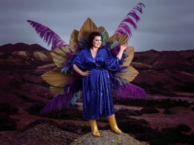 Kiri Pritchard McClean stands on a cliff in a blue sequin dress and yellow boots