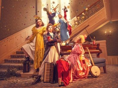 5 performers posed down an ornate carpeted staircase next to wall papered regency walls, confetti falls from the ceiling and they hold musical instruments and alcoholic drinks