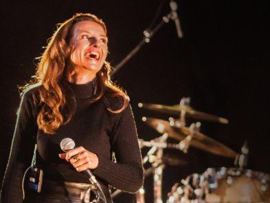 Woman dressed as Karen Carpenter with a black turtle neck jumper on, and smiling. She is holding a microphone and there is a gold drum kit behind her. 