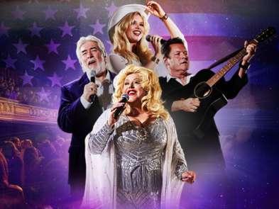 Two male and two female performers. One woman is dressed as dolly parton, another is wearing a cowboy hat. The man on the left is holding a microphone and the man on the right an acoustic guitar. The backdrop is an american flag. 