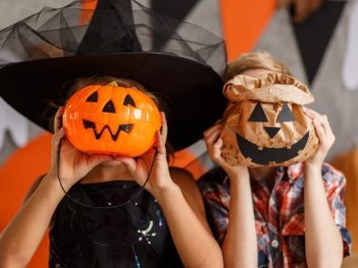 Two children holding up pumpkin themed bags and baskets in front of their faces 